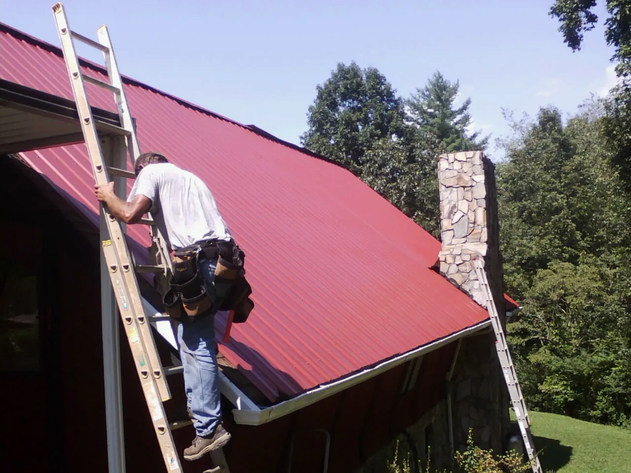 Roofer on a ladder beside a red metal roof and stone chimney — Soffit & Fascia Repair by Roofing Brothers in Rural Valley
